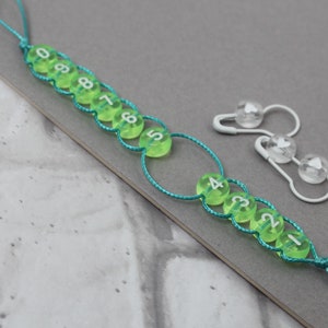 Könnte beinhalten: Eine grüne Schnur mit grünen und weißen Perlen, die von 0 bis 9 und von 1 bis 4 nummeriert sind. Zwei weiße Sicherheitsnadeln mit durchsichtigen Perlen sind ebenfalls auf dem Bild zu sehen.