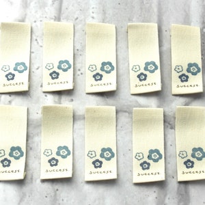 May include: Ten beige fabric tags with blue flower designs and the word "success" printed on each tag.