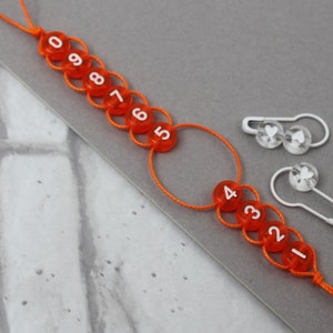 May include: Orange and white numbered stitch markers with a white string attached. Two white heart-shaped stitch markers are also included.