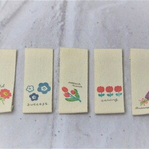 May include: Five beige fabric tags with different floral designs and text. The tags read "cheerful ness", "success", "reach dreams", "caring", and "anewassion".