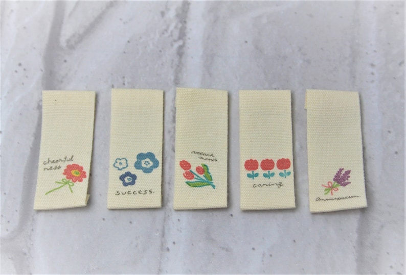 May include: Five beige fabric tags with different floral designs and text. The tags read "cheerful ness", "success", "reach dreams", "caring", and "anewassion".