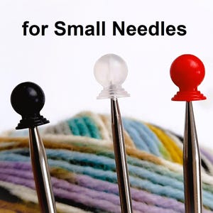 May include: Three knitting needles with different coloured plastic tips: black, clear and red. The needles are resting on a ball of colourful yarn. The text "for Small Needles" is visible above the needles.