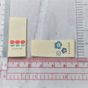 May include: Two fabric labels with different designs. The first label is cream colored with three red flowers and the text "caring". The second label is cream colored with three blue flowers and the text "success."