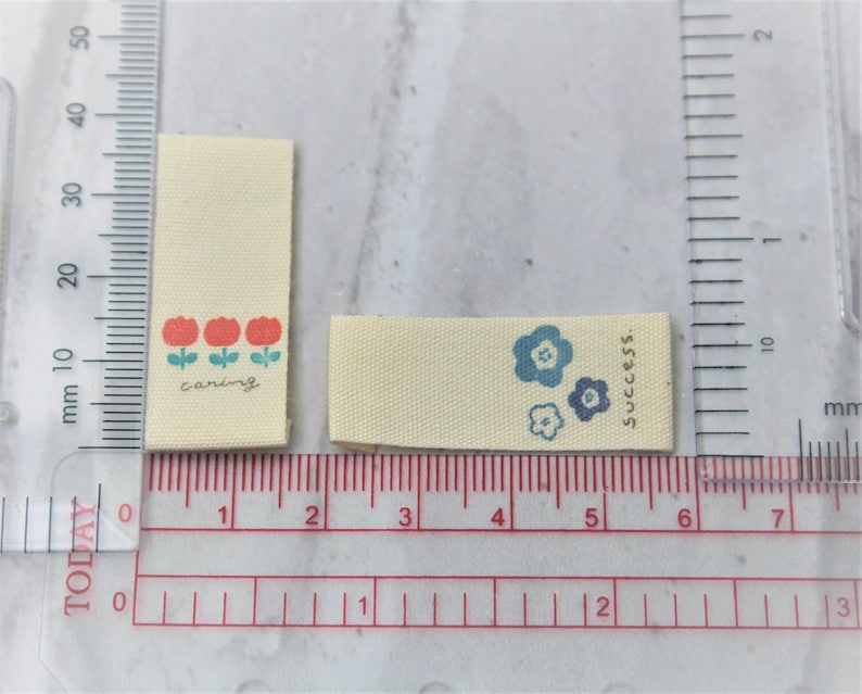 May include: Two fabric labels with different designs. The first label is cream colored with three red flowers and the text "caring". The second label is cream colored with three blue flowers and the text "success."