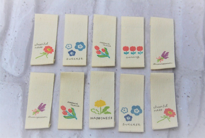 May include: Ten beige fabric tags with colorful floral designs and text. The tags feature phrases like "cheerful ness", "success", "caring", "happiness", and "anew inspiration".