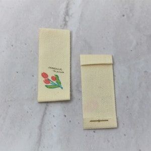 May include: Two off-white fabric labels with a small floral design and the text "attach name".