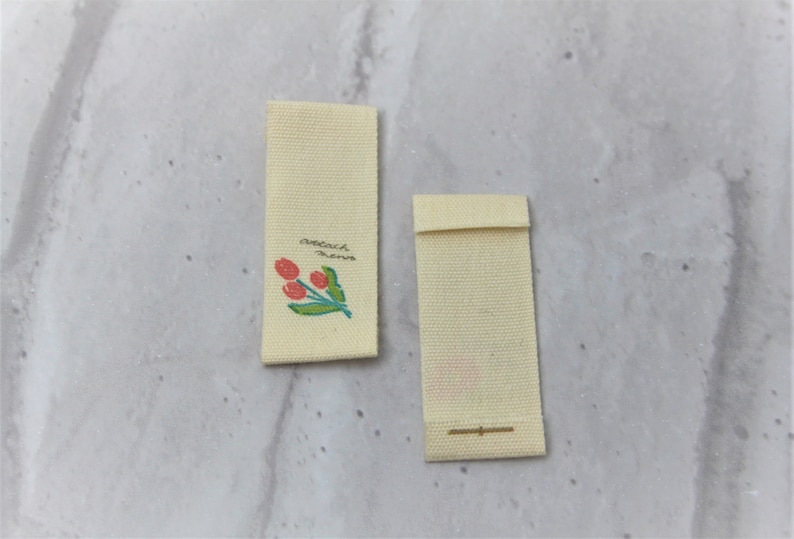May include: Two off-white fabric labels with a small floral design and the text "attach name".