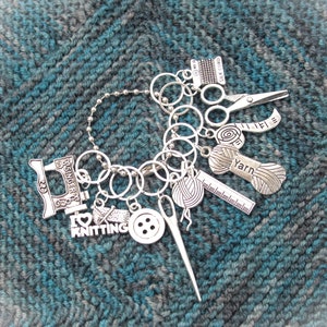 May include: A silver charm bracelet featuring a variety of knitting-themed charms, such as a sewing machine, yarn, scissors, a ruler, and a button. The charms are attached to a chain with silver rings.