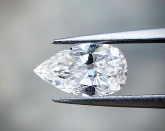 May include: A clear pear-shaped diamond held by tweezers. The diamond is faceted and reflects light.
