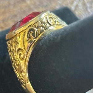 May include: A gold-colored ring with a large, faceted red gemstone. The ring features intricate, engraved floral and scrollwork designs on the band and setting. The ring is displayed against a dark background, highlighting its details.