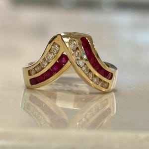 May include: A V-shaped gold ring featuring two parallel channels. One channel is set with a row of round clear diamonds, while the adjacent channel contains a row of deep red rubies. The metal band has a polished finish and a chevron design.
