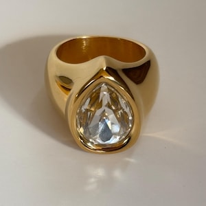 May include: A gold-toned ring with a large, clear, teardrop-shaped gemstone. The ring has a thick band and a polished finish, reflecting light. The gemstone is set in a gold bezel, creating a striking contrast. The ring is a statement piece.