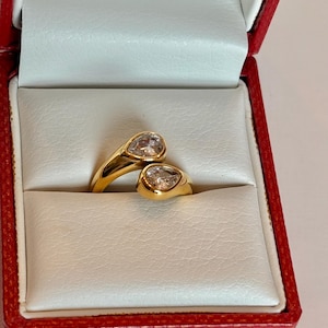 May include: A gold-colored ring with two pear-shaped clear stones. The ring is displayed in a red jewelry box with a white interior. The ring's design features a bypass style, with the band wrapping around the finger.
