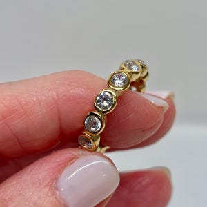 May include: A gold ring featuring a series of clear, round gemstones set in individual bezels. The ring has a polished, yellow-gold finish. The gemstones are evenly spaced around the band, creating a continuous pattern. The ring is held between fingers.