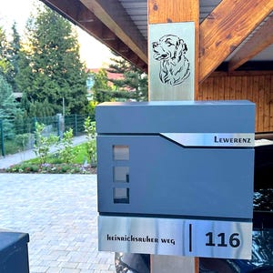 Puede incluir: Un buzón gris moderno con una placa de dirección de acero inoxidable que muestra el número "116" y el nombre de la calle "Heinrichsruher Weg". Encima, un letrero metálico presenta una silueta de perro. La palabra "LEWERENZ" está impresa en el buzón.
