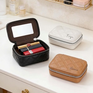 May include: Black, white, and tan quilted leather jewelry cases with silver zippers. One case is open, revealing a mirror and lipsticks. The white case is labeled "Freya", and the tan case is labeled "Aurora".
