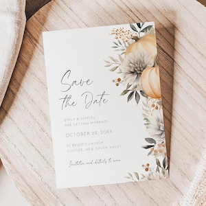Pode incluir: Um cartão de convite de casamento com o texto "Save the Date" em escrita elegante. O cartão apresenta ilustrações florais e de abóboras em tons suaves. O cartão está sobre uma superfície de madeira com um tecido de linho.