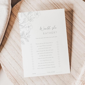 May include: A white card with the text "Would she rather?" and floral illustrations, resting on a wooden surface. The card features a list of questions. A beige linen cloth and dried plant sprig are in the background.