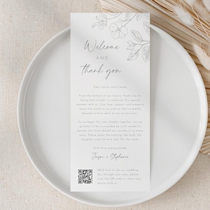 May include: A white wedding welcome card with floral accents and the text "Welcome and thank you" is displayed on a white plate. Dried, fluffy beige flowers are arranged to the right. The card includes a QR code.