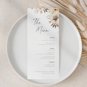 Pode incluir: Um cartão de menu branco com detalhes florais e as palavras "The Menu" em escrita elegante, repousando sobre um prato branco. O menu lista opções de entrada, pratos principais e sobremesa. Flores secas são dispostas à direita.