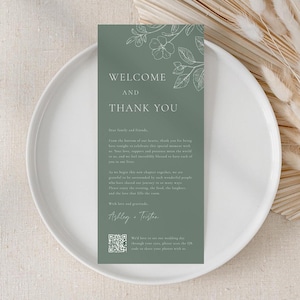 May include: A sage green wedding welcome and thank you card with floral illustrations. The card features the text "WELCOME AND THANK YOU" at the top, with additional text below. The card is placed on a white plate, with dried flowers in the background.