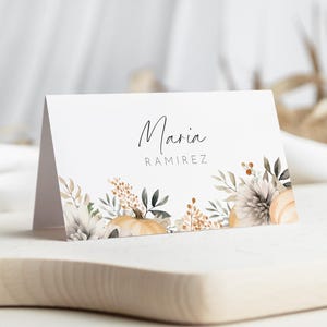 Pode incluir: Um cartão de lugar branco dobrado com o nome "Maria" e o sobrenome "Ramirez" em escrita elegante. O cartão é decorado com abóboras, flores e folhagens em aquarela em tons de creme, bege e verde.