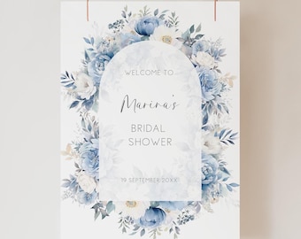 Puede incluir: Un letrero blanco con un diseño floral y el texto "Welcome to Marina's Bridal Shower". El arreglo floral presenta flores y follaje azules y blancos. El letrero está suspendido por dos finos cordones marrones.