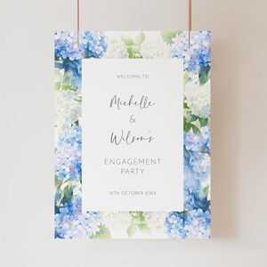 May include: A rectangular engagement party sign with a floral border of blue and white hydrangeas. The sign reads "Welcome to Michelle & Wilson's Engagement Party" with the date "19th October 20XX" at the bottom.