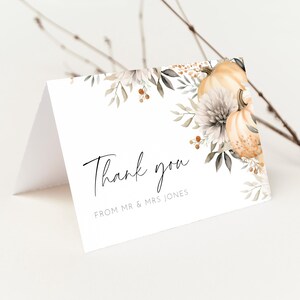 Pode incluir: Um cartão de agradecimento branco dobrado com um design floral em aquarela com abóboras e folhagens. As palavras "Thank you" estão escritas em um script elegante, com "FROM MR & MRS JONES" abaixo. Um galho está no fundo.