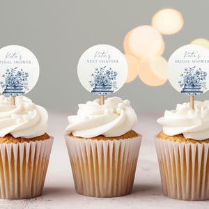 Peut inclure: Trois cupcakes avec glaçage blanc et décorations. Les décorations sont rondes avec des motifs floraux bleus et le texte "Katie's Bridal Shower" et "Next Chapter". Les cupcakes sont dans des caissettes en papier.