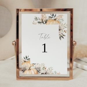 Pode incluir: Um cartão de número de mesa em uma moldura dourada. O cartão apresenta a palavra "Table" acima do número "1" em preto. Ilustrações florais em aquarela de abóboras e folhagens em tons de branco, bege e verde decoram os cantos.