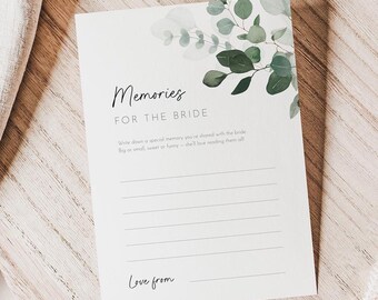May include: A white card with the text "Memories for the Bride" and lined spaces for writing. The card is decorated with green eucalyptus leaves and sits on a wooden charger. A linen napkin and dried flowers are also in the image.