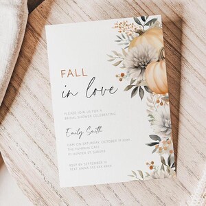 Pode incluir: Um cartão de convite branco com o texto "FALL in love" e ilustrações florais e de abóboras. O cartão está sobre uma superfície de madeira com um pano de linho e plantas secas.