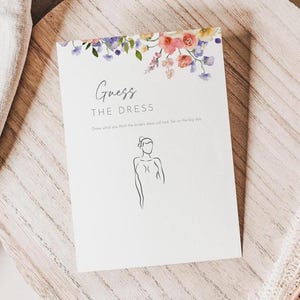 Puede incluir: Una tarjeta blanca con el texto "Guess the Dress" y un dibujo lineal de una mujer. La parte superior de la tarjeta está decorada con flores de acuarela en rosa, morado y azul. La tarjeta está sobre una superficie de madera.