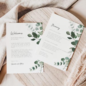 May include: Two white rectangular cards featuring green eucalyptus leaf illustrations. The left card is titled Welcome and the right card is titled Itinerary, both containing printed text details for a wedding weekend event in black ink.