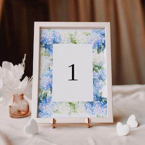 May include: White frame with a floral design featuring blue and white hydrangeas. The frame holds a white card with the number "1" printed in black.