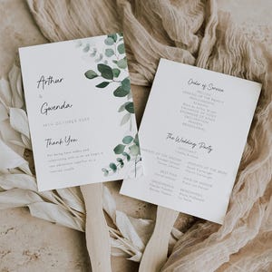 Könnte beinhalten: Zwei weiße Hochzeitsfächer mit Holzgriffen. Ein Fächer zeigt die Namen "Arthur & Gwenda" und das Datum "19. Oktober 20XX" mit Eukalyptusblatt-Akzenten. Der andere Fächer listet die Reihenfolge der Zeremonie und die Details der Hochzeitsgesellschaft auf.