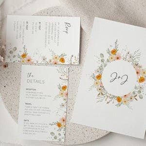 May include: Wedding invitation suite with a floral design. The invitation features a wreath of orange and white flowers with the text "J+J" in the center. The RSVP card includes details for attending by text or post.