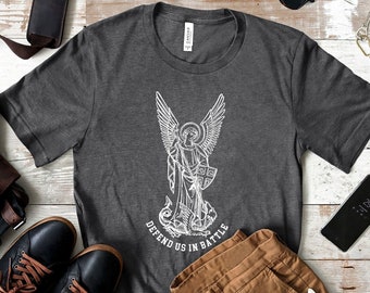 May include: A dark gray t-shirt with a white graphic of an angel with wings and a sword, standing on a dragon. The text "Defend Us In Battle" is printed below the graphic.