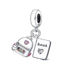 Puede incluir: Un charm de plata con un colgante de mochila y otro de libro. La mochila tiene un corazón rosa, una cara sonriente y piedras de colores. El colgante rectangular de libro tiene grabado el texto book y una piedra clara en forma de corazón. S925.