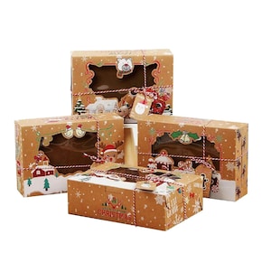 Puede incluir: Cuatro cajas de regalo rectangulares de cartón con ventanas transparentes, decoradas con ilustraciones navideñas que incluyen renos, muñecos de nieve y casas. Cada caja está atada con cordel rojo y blanco y tiene una pequeña etiqueta colgante.
