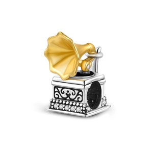 May include: A silver charm in the shape of a vintage record player with a gold-toned horn. The charm is decorated with clear cubic zirconia stones.