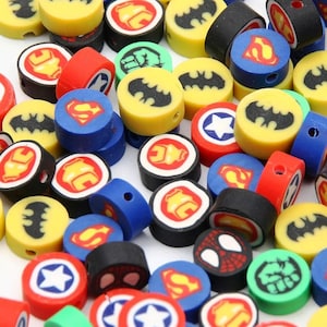 Op de afbeelding: Een verzameling kleine, ronde, veelkleurige kralen met diverse superheldenlogo's en symbolen, waaronder het Batman-vleermuisembleem, het Superman S-schild, het Spider-Man-masker, de Captain America-ster en de Hulk-vuist, elk voorzien van een gat in het midden.