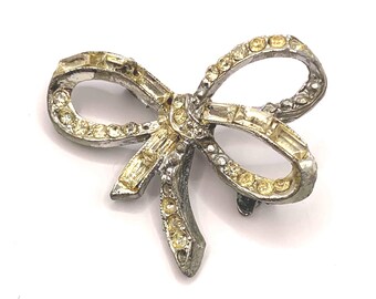 May include: A silver bow brooch with clear and yellow rhinestones. The bow is tied in a knot with the ends of the bow trailing down.