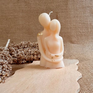 May include: A pair of candle sculptures in the shape of two people embracing. The candle is a light beige color with gold accents.