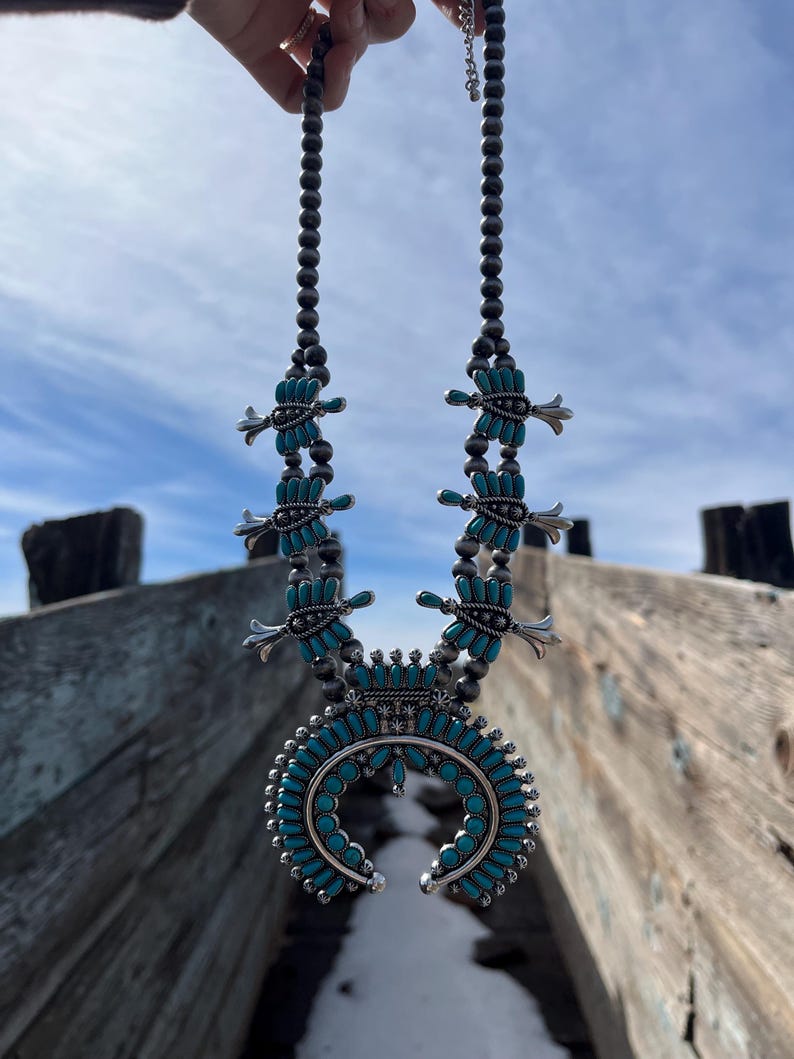 May include: A turquoise and silver Native American style necklace with a crescent moon pendant. The necklace is made of small turquoise beads and silver beads. The pendant is a crescent moon shape with a turquoise inlay.