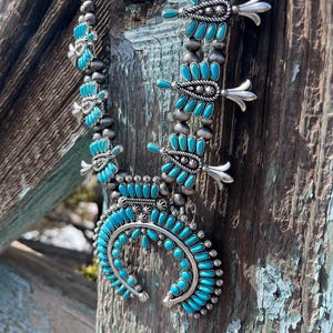 May include: A turquoise and silver Native American necklace with a crescent moon pendant and feather designs. The necklace is hanging on a weathered wooden surface.