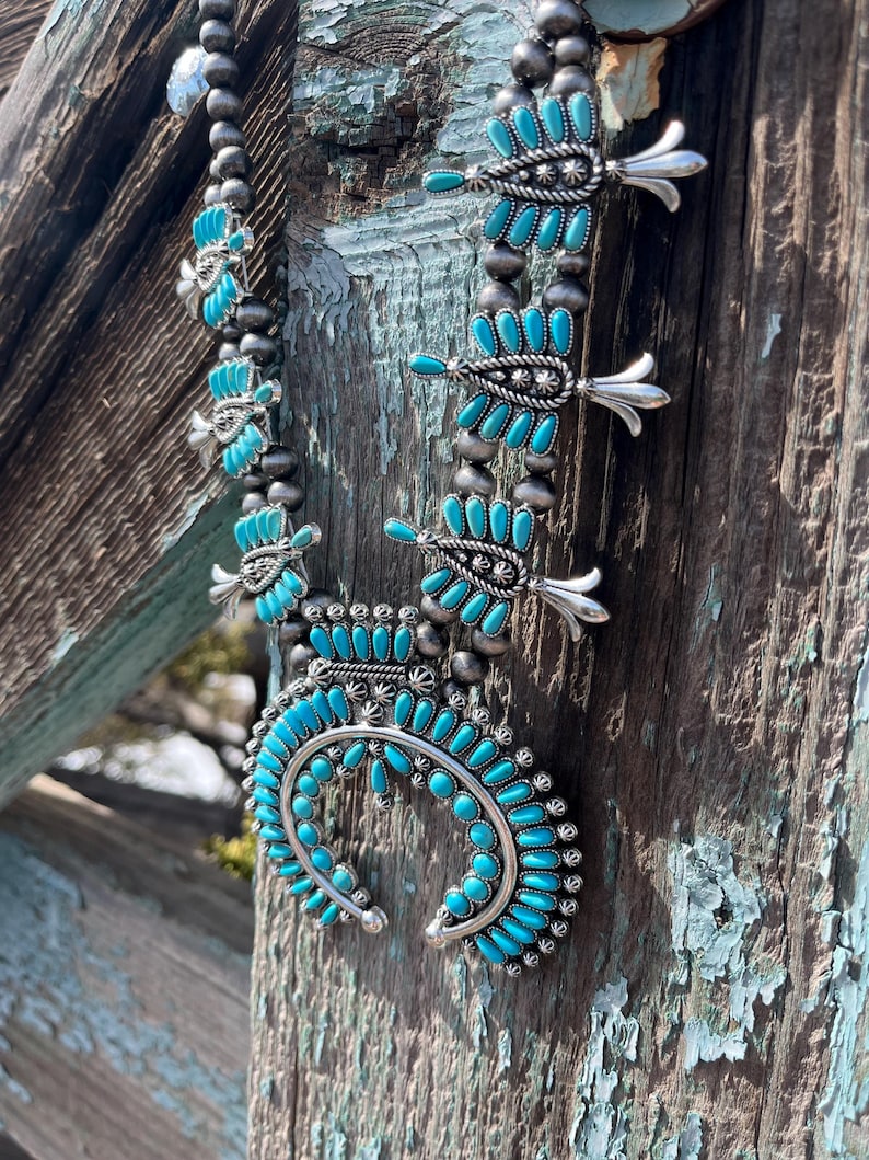 May include: A turquoise and silver Native American necklace with a crescent moon pendant and feather designs. The necklace is hanging on a weathered wooden surface.