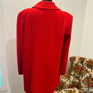 May include: A bright red coat displayed on a black mannequin. The coat has a classic design with a notched collar and long sleeves. The coat is made of a smooth, solid fabric. The background includes a floral patterned chair.