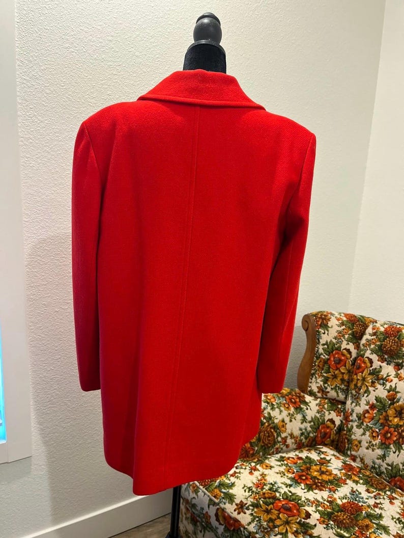 May include: A bright red coat displayed on a black mannequin. The coat has a classic design with a notched collar and long sleeves. The coat is made of a smooth, solid fabric. The background includes a floral patterned chair.